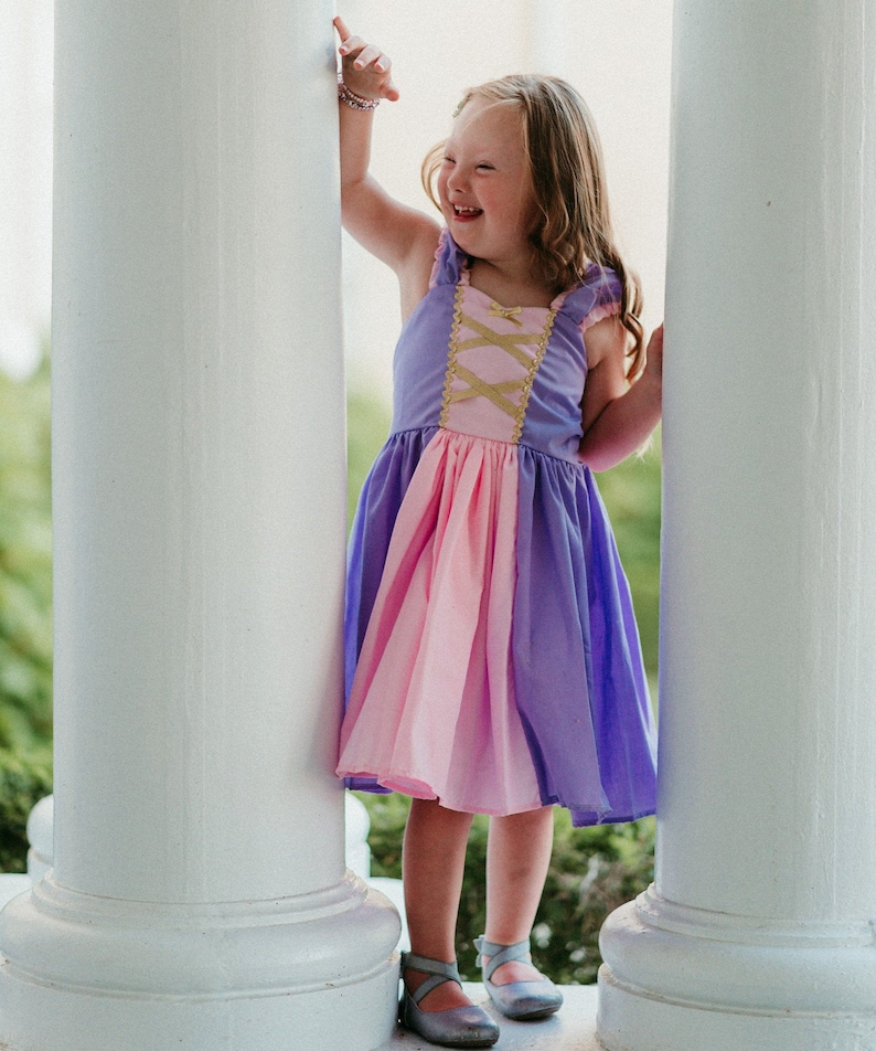 May include: A young girl wearing a purple, pink, and gold dress, standing between two white pillars. The dress has a bodice with criss-cross gold detailing and a layered skirt. She is smiling and wearing silver shoes.