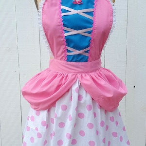 May include: A pink and blue apron with a white lace trim and pink polka dots. The apron has a heart-shaped bodice with a criss-cross ribbon detail.