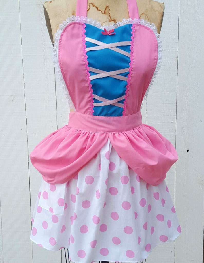 May include: A pink and blue apron with a white lace trim and pink polka dots. The apron has a heart-shaped bodice with a criss-cross ribbon detail.