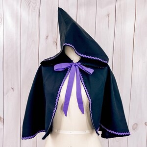 May include: A black hooded cape with a purple trim and a purple ribbon tied at the neck. The cape is on a white dress form.