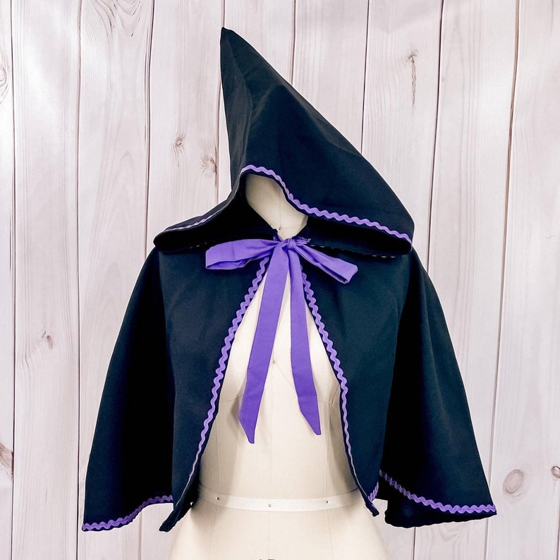 May include: A black hooded cape with a purple trim and a purple ribbon tied at the neck. The cape is on a white dress form.