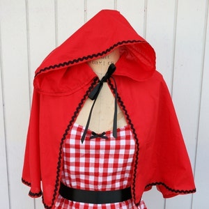 May include: Red hooded cape with black trim and a black ribbon tied at the neck. The cape is worn over a red and white gingham dress with a black belt.