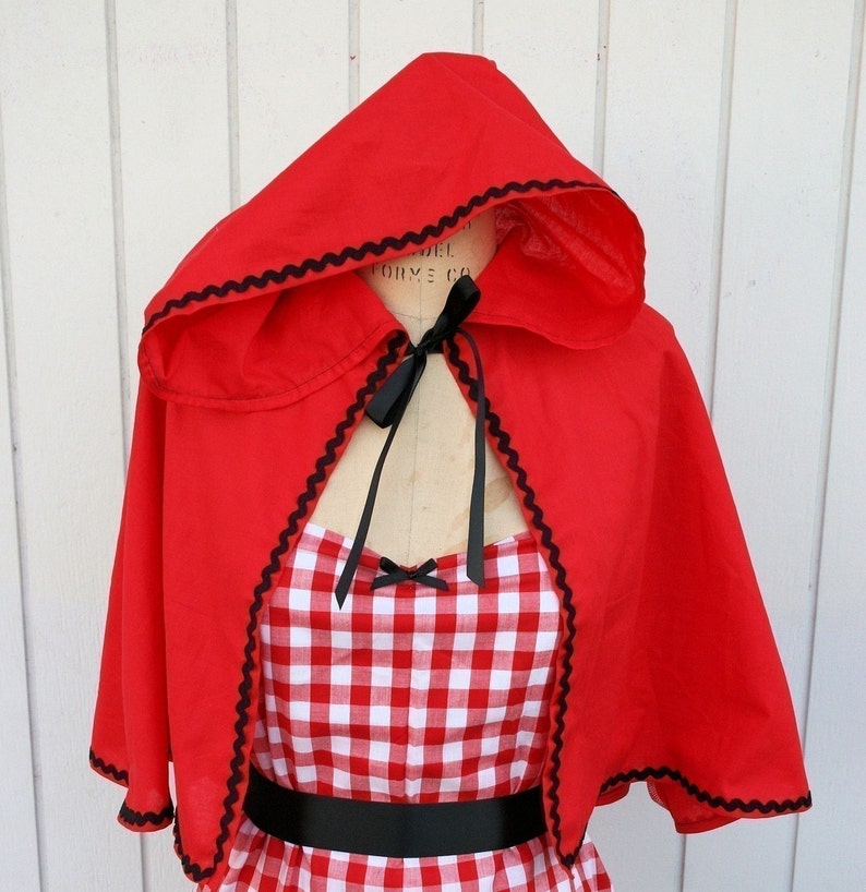 May include: Red hooded cape with black trim and a black ribbon tied at the neck. The cape is worn over a red and white gingham dress with a black belt.