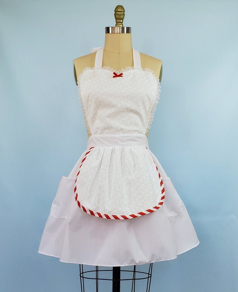 May include: White apron with a red and white striped trim around the bottom edge. The apron has a heart-shaped top with a red bow and a gathered skirt.
