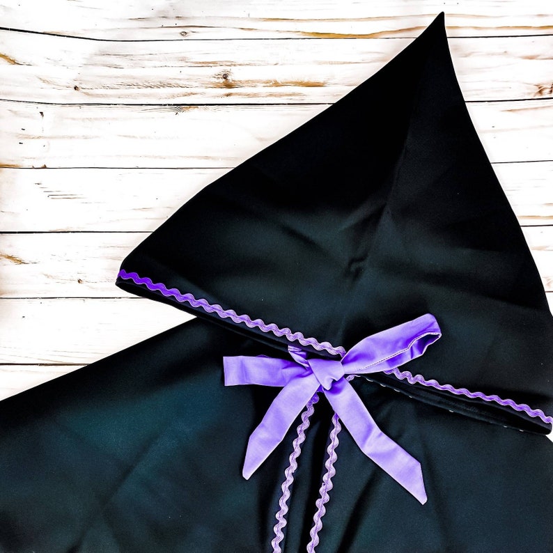 May include: A black hooded cape with a purple satin ribbon tied in a bow at the neck. The cape has a scalloped edge.