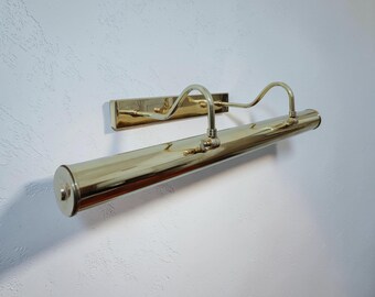 May include: A brass picture light fixture mounted on a white wall. The light has a long, cylindrical body with a curved arm and a rectangular base. The fixture is made of shiny brass.