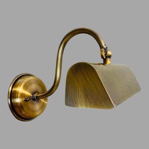 May include: A wall-mounted brass lamp with a curved arm and a rectangular shade. The lamp has a circular base and a vintage aesthetic. The brass finish gives the lamp a warm, antique look, suitable for interior lighting.