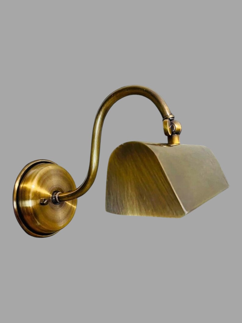 May include: A wall-mounted brass lamp with a curved arm and a rectangular shade. The lamp has a circular base and a vintage aesthetic. The brass finish gives the lamp a warm, antique look, suitable for interior lighting.
