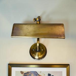 May include: A vintage-style brass wall-mounted lamp with a rectangular shade and a round base. The lamp has a classic design, with a warm, golden hue. The lamp is mounted above a framed picture.