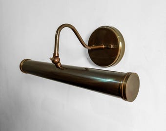 May include: A vintage brass picture light fixture. The fixture has a cylindrical body, a curved arm, and a circular wall mount. The brass finish gives it a classic, antique look. The light is designed to illuminate artwork or other wall-mounted items.