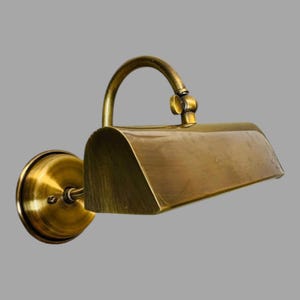 May include: A brass-toned wall-mounted lamp with a curved arm and a rectangular shade. The lamp has a circular base and a classic, vintage design. The finish is a warm, antique brass color, suitable for interior lighting.