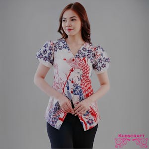 Könnte beinhalten: Eine weiße Bluse mit V-Ausschnitt und kurzen Puffärmeln. Die Bluse hat ein lebendiges Pfauenmuster in Rot, Blau und Weiß mit floralen Akzenten. Der untere Teil der Bluse ist offen. Der Markenname "KUDSCRAFT" ist unten rechts sichtbar.