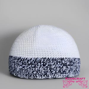 May include: White and navy blue kufi hat. The top of the hat is white, with a textured pattern. The bottom band is navy blue with a geometric design. The hat is dome-shaped and sits on a gray background.