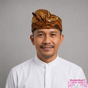 May include: A traditional Balinese udeng headpiece featuring a golden brown batik fabric with intricate geometric and floral patterns. The headgear is tied with a prominent knot at the top and displays a structured, folded design with a dark brown base.