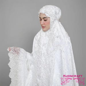 May include: A white prayer garment featuring delicate lace embroidery. The head covering and shawl are decorated with floral designs. The fabric appears soft and flowing. The brand name "KUDSCRAFT" is displayed in the bottom right corner.
