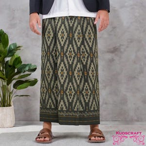 Puede incluir: Un sarong largo estampado en tonos verde oliva, beige y naranja. La tela presenta un diseño geométrico repetitivo. El borde inferior tiene un borde verde oscuro con detalles dorados. El sarong se usa con sandalias marrones.