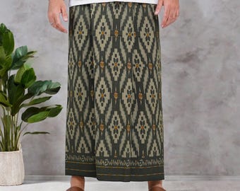 May include: A sarong featuring a dark olive green and black geometric ikat pattern with repeating diamond shapes and small orange accents. The fabric has a horizontal border at the hem with intricate line details. The garment is worn with a white shirt.