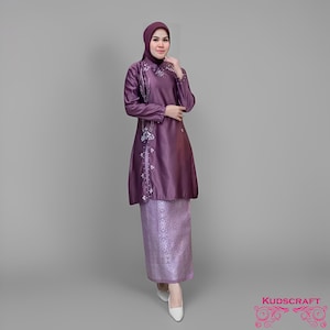 Puede incluir: Un conjunto morado de dos piezas con un velo a juego. La parte superior de manga larga presenta detalles florales bordados y un corte tipo túnica. La falda es de un tono morado a juego con un patrón sutil. Zapatos blancos completan el conjunto.