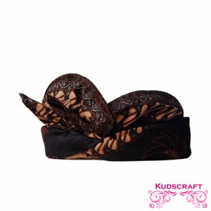 Puede incluir: Un tocado tradicional con un estampado batik marrón y negro. El tocado presenta un diseño plegado con detalles intrincados y un motivo floral decorativo. El producto es de Kudscraft.