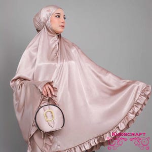 May include: A light pink satin prayer shawl with a ruffled trim. The shawl is draped over a person's shoulders and has a matching head covering. A small round purse with a gold buckle is held in the person's hand.