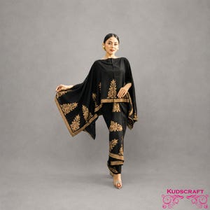 May include: A black ensemble featuring a cape and matching pants, adorned with gold floral embroidery. The cape drapes over the shoulders, and the pants are tailored. The outfit is set against a gray backdrop, showcasing the intricate details of the design.