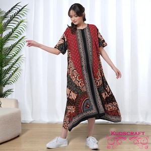 Puede incluir: Vestido hasta la rodilla en rojo, negro y marrón con estampado. Tiene mangas cortas y un panel central negro. El vestido presenta bordes decorativos y un corte holgado. La marca "KUDSCRAFT" es visible.