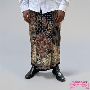 May include: A patterned sarong in black, brown, and gold, with floral and geometric designs. The sarong is worn with a white shirt and black boots. The image is taken from the waist down.