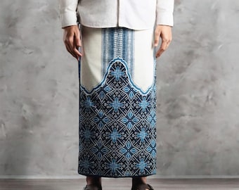 May include: A long, patterned sarong in shades of blue and white. The lower portion features a detailed geometric design, while the upper section has vertical stripes. The sarong is worn by a person wearing a white shirt and sandals.