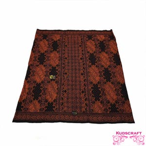 May include: A rectangular batik textile with a repeating pattern in shades of rust and black. The design features floral and geometric shapes. A small, gold-colored clasp is visible near the center. The textile is displayed on a white background.