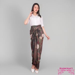 Op de afbeelding: Een vrouw draagt een wit overhemd en een lange, bedrukte rok. De rok heeft een donkere achtergrond met bloemen en geometrische patronen in bruin, groen en roze. Het is gewikkeld en vastgemaakt met een decoratieve gesp.