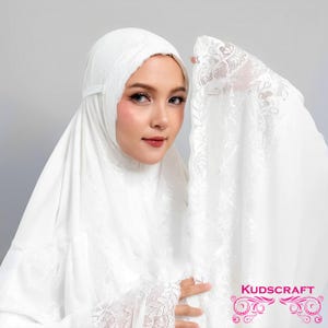 May include: White prayer shawl with lace trim. The shawl is draped over a person's head and shoulders. The word "KUDSCRAFT" is printed on the shawl.