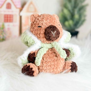 May include: A handmade, plush bear in shades of brown, with a dark brown snout and paws. The bear wears a light green and white scarf. The bear is sitting on a white, fluffy surface. The bear is a cozy, handmade item.