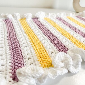 May include: A crocheted baby blanket with a striped pattern in white, yellow, and purple. The blanket has a ruffled edge.