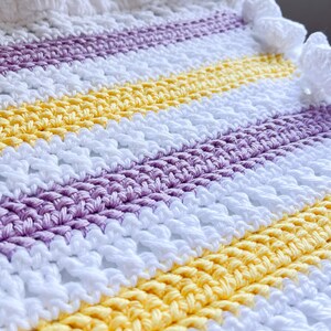 May include: A crocheted baby blanket with a striped pattern in white, yellow, and purple. The blanket has a white crocheted border with pom-poms.