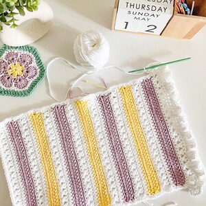 May include: A white crocheted blanket with stripes of yellow and purple yarn. The blanket has a scalloped edge.