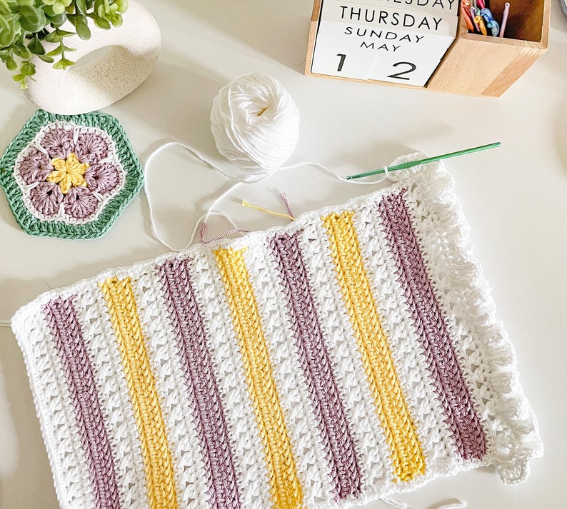 May include: A white crocheted blanket with stripes of yellow and purple yarn. The blanket has a scalloped edge.