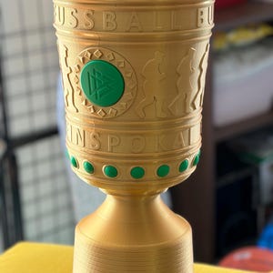 Op de afbeelding: Een goudkleurige trofee met de tekst FUSSBALL BUNDESPOKAL, reliëf-silhouetten van rennende figuren en een centraal groen rond embleem met een geometrisch ontwerp. De rand en het onderste gedeelte zijn versierd met afwisselend groene en witte stippen.