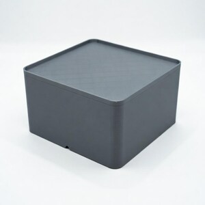 May include: A gray, square storage box with a matching lid. The box has rounded corners and a smooth, matte finish. The lid sits flush on top of the box, creating a clean, minimalist look. The box is made of a solid material and is suitable for various storage needs.