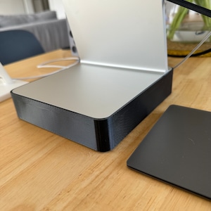 May include: A silver and black computer stand with a black square accessory. The stand has a rectangular base and a vertical support for a monitor. The accessory is a small, square, black device. The stand is on a wooden surface.