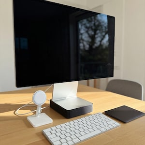 May include: A modern computer setup featuring a large black monitor, a white keyboard, and a black trackpad on a light wood desk. A white charging stand and a black and white speaker are also present, creating a clean, minimalist aesthetic.