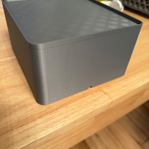 Puede incluir: Una caja rectangular gris de impresión 3D con un acabado liso y mate. La caja está sobre una superficie de madera clara.