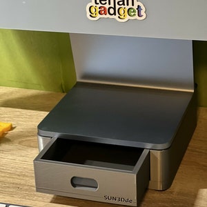 Könnte beinhalten: Ein grauer 3D-gedruckter Ständer mit einer Schublade, die herausgezogen werden kann. Der Ständer hat eine schwarze Oberseite und die Schublade ist mit "SUN3Dde" beschriftet.
