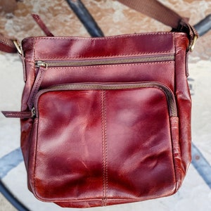 May include: A burgundy leather crossbody bag with multiple zipped compartments. The bag has a flap closure and a long adjustable strap.