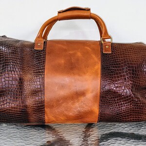May include: A brown leather duffel bag with a crocodile-embossed design. The bag has a top handle and a shoulder strap.