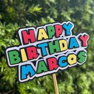May include: A colorful cake topper with the words "Happy Birthday Marcos" in a bold, blocky font. The letters are outlined in black and have a white background.
