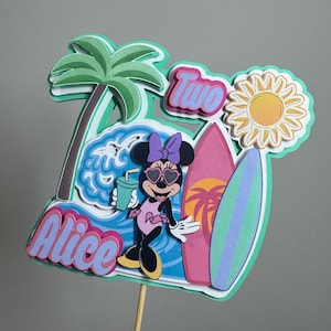 May include: A colorful cake topper featuring Minnie Mouse in sunglasses, holding a drink, and standing next to surfboards. The topper includes the name "Alice" and the number "Two" in a beach-themed design with a palm tree and sun.