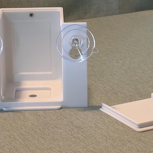 Puede incluir: Un soporte de ducha de pared de plástico blanco con dos ventosas transparentes en los laterales para su fijación. La unidad tiene un compartimento rectangular abierto con un pequeño orificio de drenaje en la base e incluye una tapa blanca a juego.