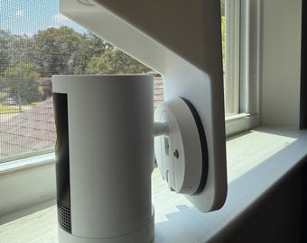 Puede incluir: Un sistema de cámara de seguridad blanco montado en un alféizar. El sistema incluye una unidad de cámara cilíndrica y un soporte de montaje rectangular. La cámara está posicionada para capturar vistas exteriores, con una pantalla y árboles visibles a través de la ventana.