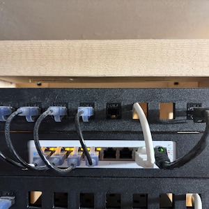 May include: Close-up of a network switch and patch panel with multiple Ethernet cables connected. The switch is white with several ports, and the panel is black. The cables are various colors, including blue and black, and are neatly organized.