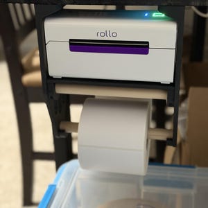 Op de afbeelding: Een witte en paarse Rollo-etikettenprinter is onder een donkere houten tafel gemonteerd. Een rol witte etiketten wordt onder de printer gehouden. Op de printer staat het woord "rollo".